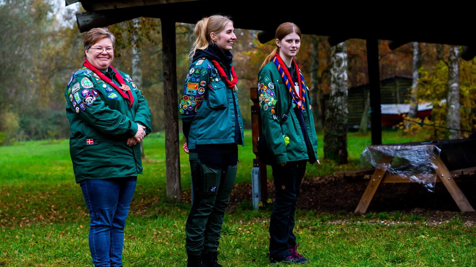 Tre spejdereledere stå udenfor, en kigger direkte ind i kamaraet.